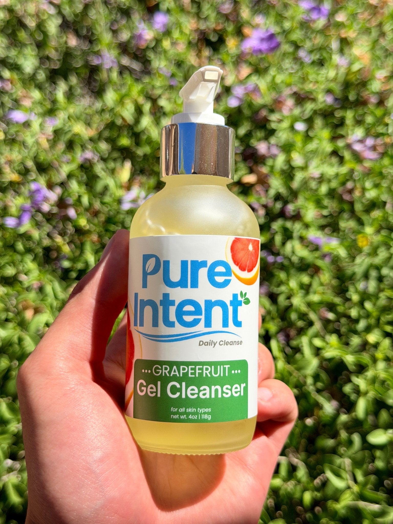 Hand holding a bottle of Pure Intent Grapefruit Gel Cleanser against a green background with flowers.